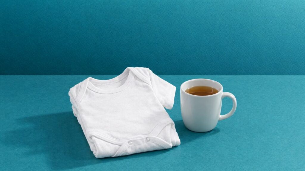 A folded white baby bodysuit beside a mug of tea