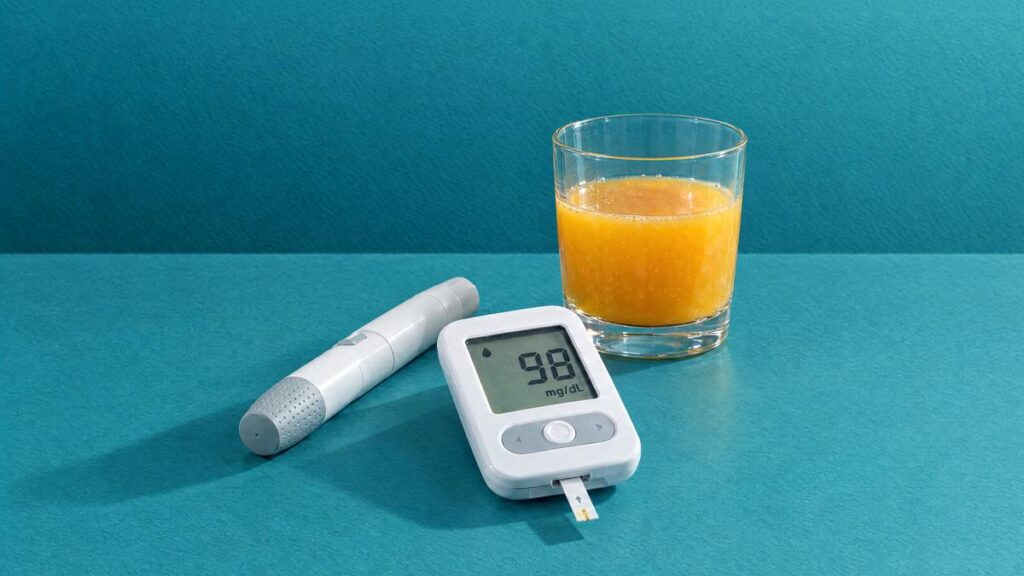 A blood glucose meter reading 98 mg/dL beside a lancing device and a glass of orange juice