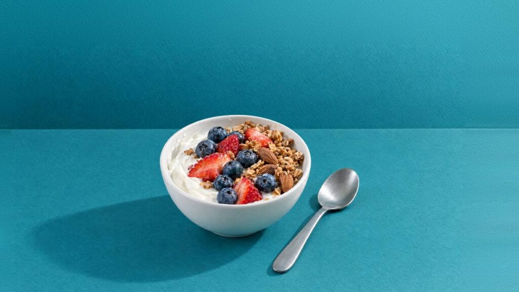 A bowl of Greek yogurt with blueberries, strawberries, granola and almonds, with a spoon beside it